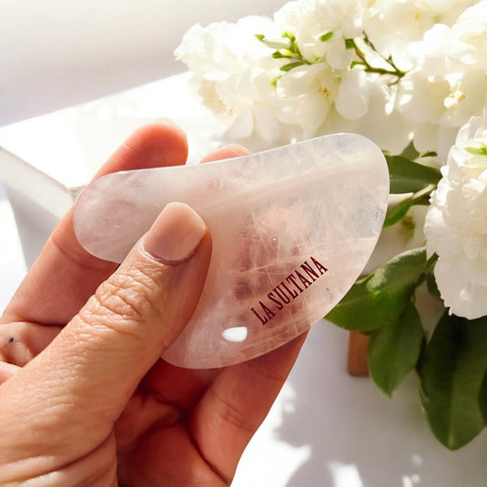 Une main tient un outil de massage facial en pierre translucide étiqueté « Ensemble GLOW Gua Sha & huile GlÞd », fabriqué à partir de quartz rose exquis, sur fond de fleurs blanches. La scÚne est lumineuse et bien éclairée, évoquant un environnement serein et propre. Ce produit est de La Sultana Savonnerie | Savons et soins naturels faits main | Québec.