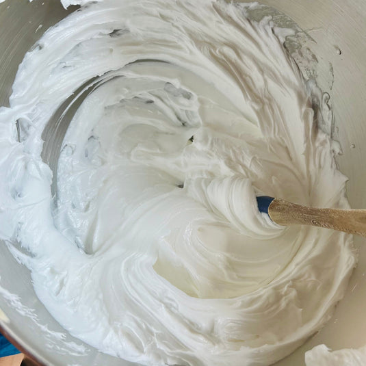 Un bol à mélanger contient un mélange succulent et épais qui est agité avec une cuillère en bois. La texture onctueuse et brillante, qui rappelle le savon fouetté FAIROUZ agrûmes sucrés de La Sultana Savonnerie | Savons et soins naturels faits main | Le Québec présente des motifs tourbillonnants à sa surface.